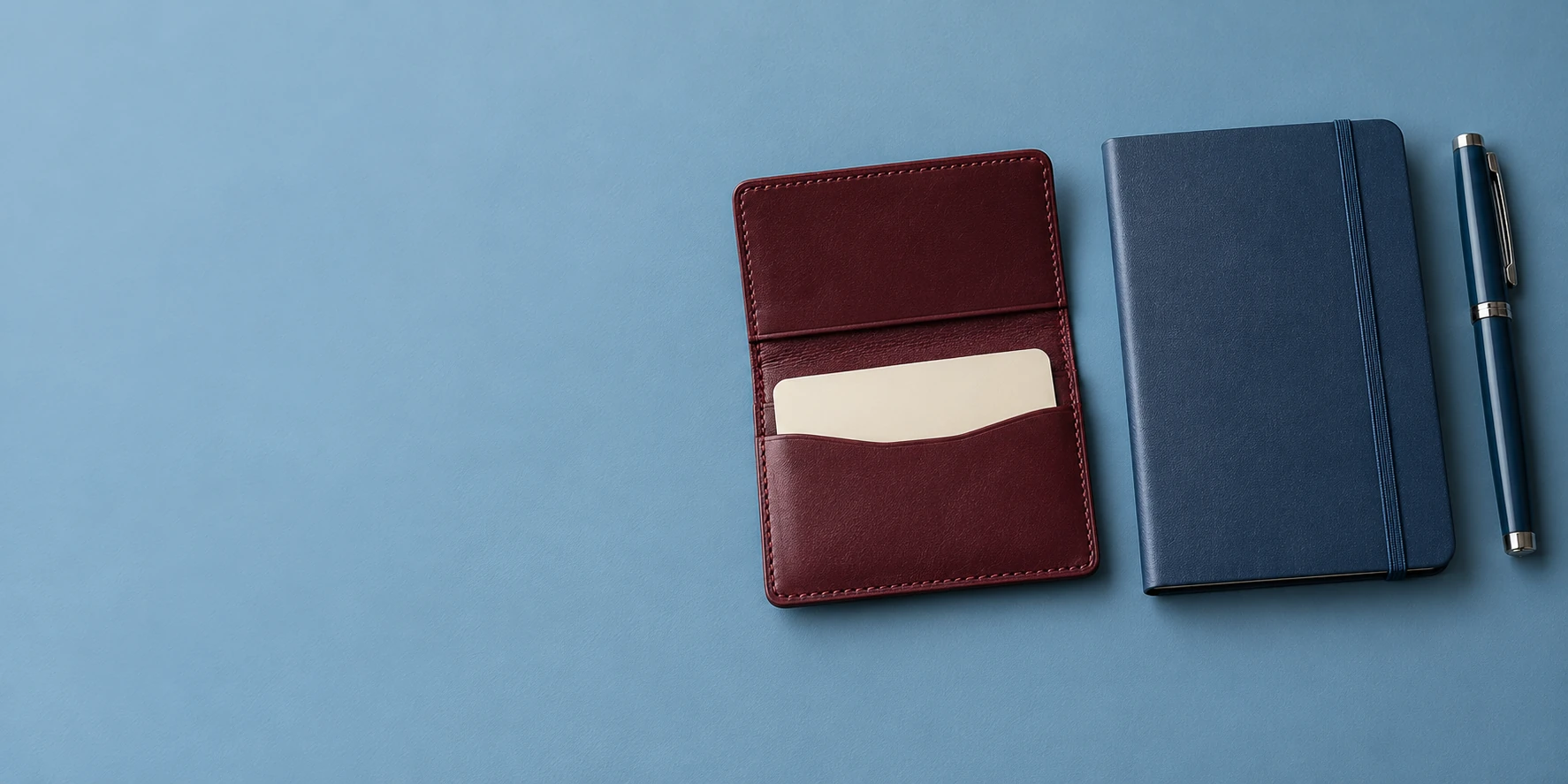 A burgundy card wallet with a blank card, a navy notebook, and a pen resting on a dusty-blue surface