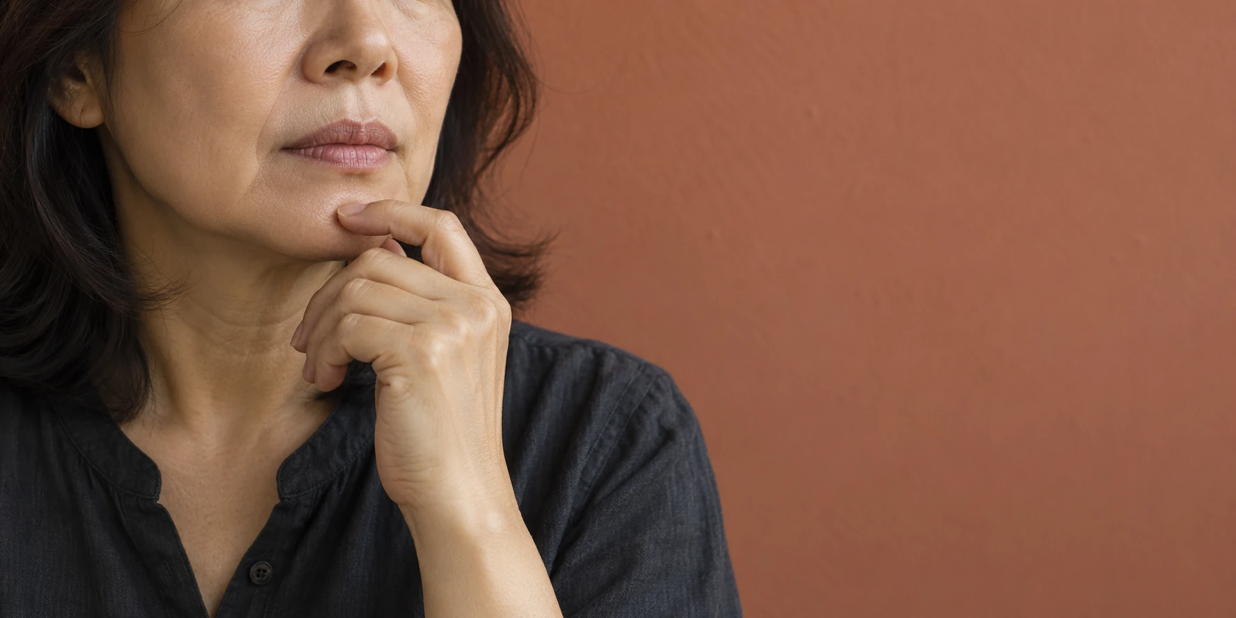 A middle-aged woman resting a hand lightly against her chin in front of a muted terracotta wall